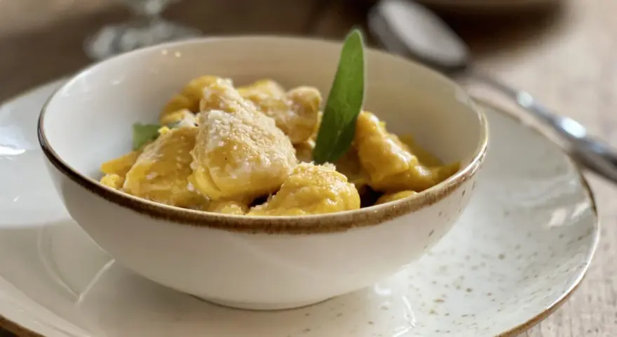 Gnocchi di zucca dorati e soffici, conditi con parmigiano e una foglia di salvia, serviti in una ciotola bianca su un piatto maculato.