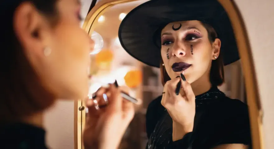 Frau mit Hexenhut und Halloween-Make-up trägt Lippenstift vor einem Spiegel auf.
