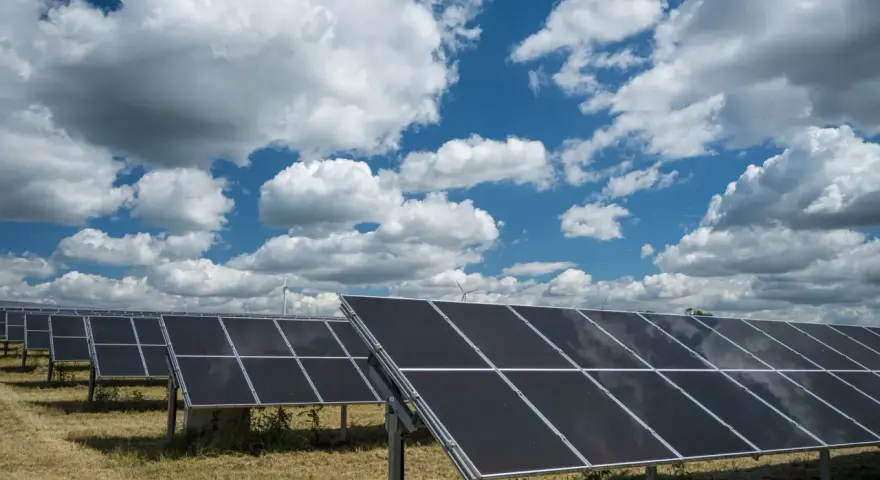 Rzędy paneli fotowoltaicznych 50kW na tle błękitnego nieba z białymi chmurami. W oddali widać turbiny wiatrowe.