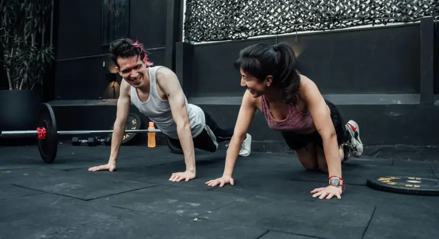 Pareja sonríe haciendo flexiones, un ejemplo de cuales son los ejercicios de fuerza.