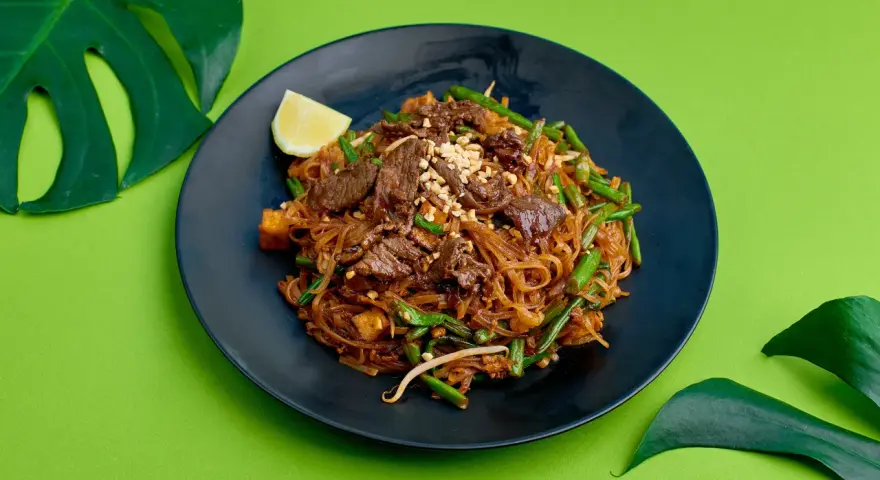 Pyszne pad thai z wołowiną, fasolką szparagową i orzeszkami na czarnym talerzu, ozdobione ćwiartką cytryny i liśćmi monstery.