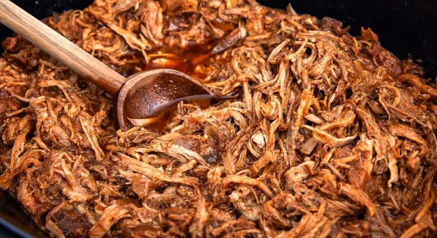 En stor gryta med saftig, långkokt pulled pork i en mörk sås. En träsked vilar i den läckra pulled pork grytan.