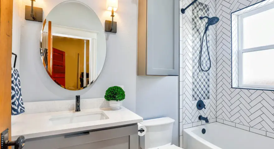 Une petite salle de bain moderne avec baignoire, lavabo gris, miroir rond et carrelage chevrons. Une belle idée pour optimiser l'espace.