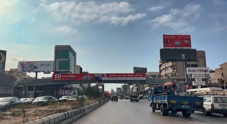 Driving in Lebanon, a busy street with cars, trucks, and billboards advertising various services and products.