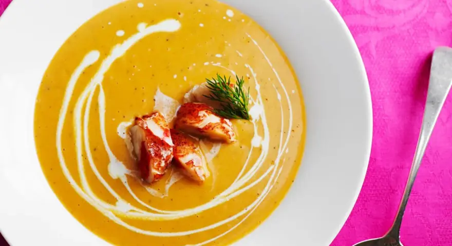 Un cremoso bisque de langosta, adornado con trozos de carne de langosta y un toque de eneldo, servido en un plato blanco sobre un mantel rosa vibrante.