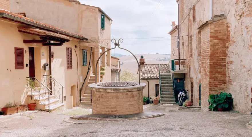 Un antico pozzo in mattoni domina una piazzetta di San Giovanni d'Asso, circondata da case in pietra e con dolci colline sullo sfondo.