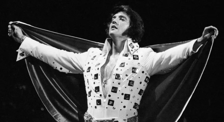 Elvis Presley, der unzählige Tonträger verkaufte, posiert in einem glitzernden weißen Anzug mit ausgebreitetem Umhang.