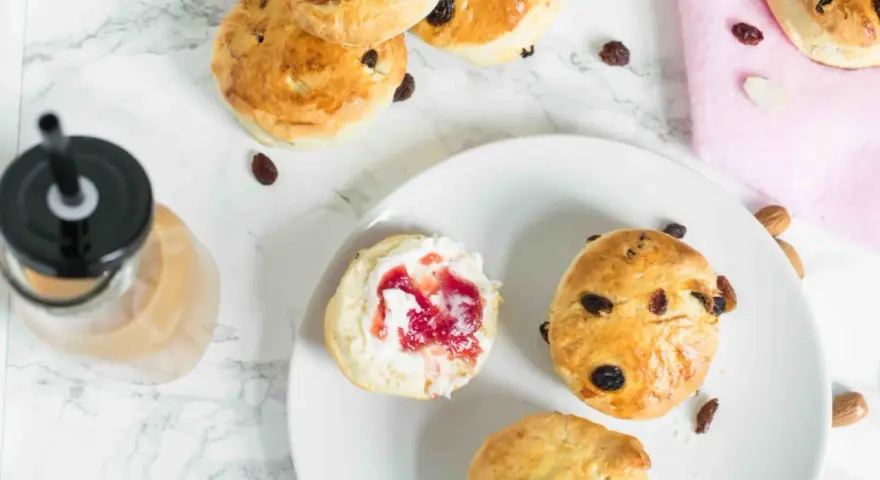 Goldbraune Scones mit Rosinen und Mandeln, ein Scones Teig mit Marmelade und Sahne, daneben ein Getränk.