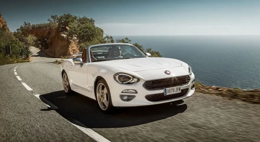 Biały Fiat 124 Spider sunie po krętej drodze nad morzem.