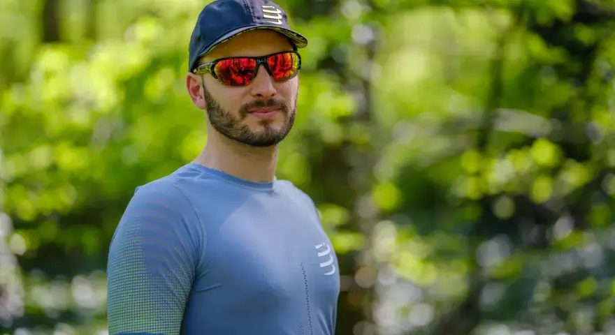 Mężczyzna w czapce i sportowych okularach przeciwsłonecznych, z odbiciem lasu w soczewkach.