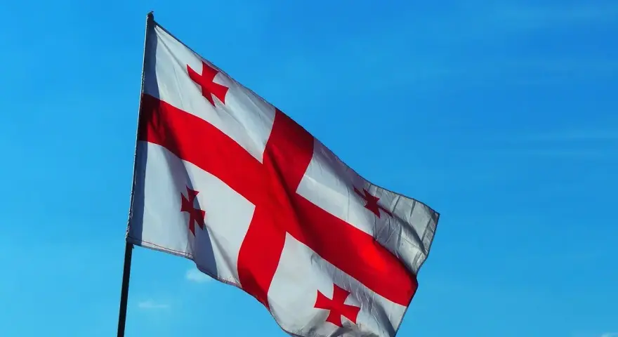 Flaga Gruzji powiewa na tle błękitnego nieba. Białe tło przecina czerwony krzyż, a w każdym z czterech pól znajduje się mniejszy czerwony krzyżyk.