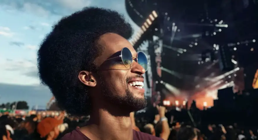 Un homme souriant avec des lunettes de soleil et des bouchons d'oreilles, profitant d'un concert en plein air. Le port des bouchons d'oreilles est obligatoire pour protéger son audition.