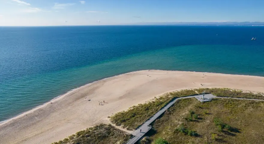 Piękne wydmy helskie nad turkusowym morzem. Drewniana kładka prowadzi przez zarośla na piaszczystą plażę, gdzie spacerują nieliczni turyści.