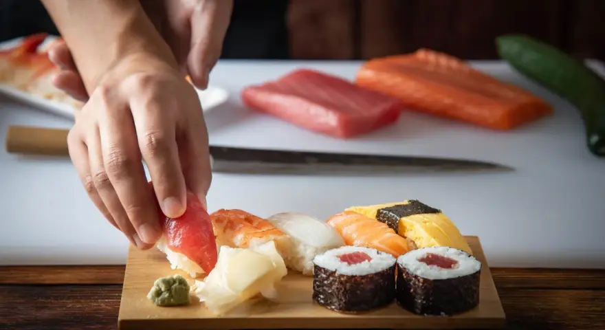 Ręce układają kawałek tuńczyka na ryżu, tworząc sushi. Na desce widać też krewetkę, kalmara, łososia i tamago. To inspiracja, jak zrobić sushi.