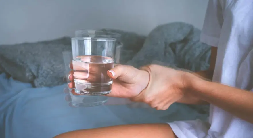Dłoń z drżącym szkłem wody, sugerującym zwidy po alkoholu. Rozmyte kontury i niepewny chwyt.