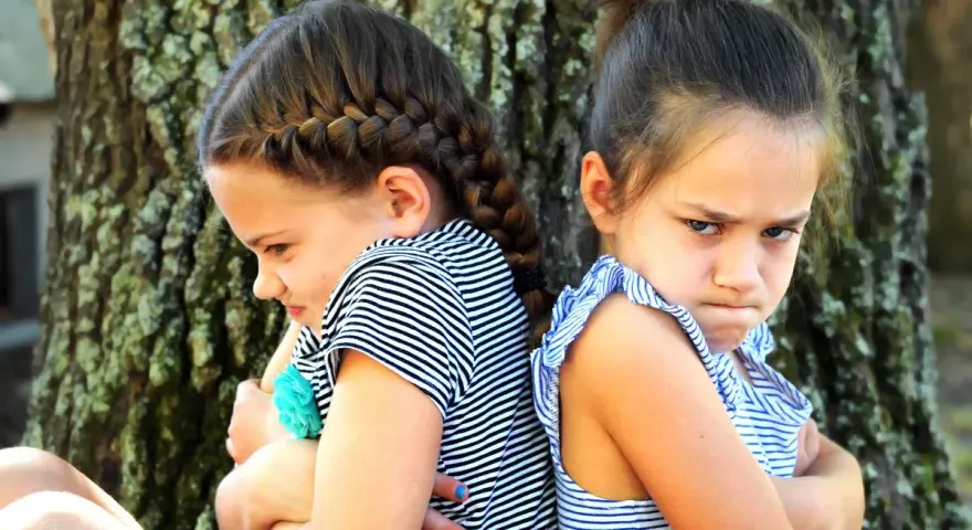 Dwie siostry siedzą plecami do siebie, z ramionami skrzyżowanymi i obrażonymi minami. Może to być odpowiedź na pytanie, dlaczego rodzeństwo się nie lubi.