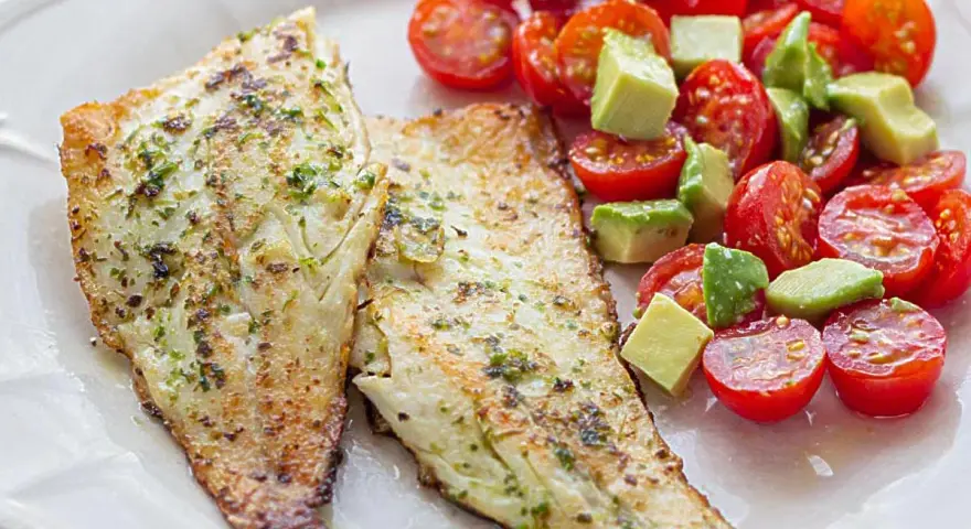 Dos filetes de lubina a la plancha con hierbas, acompañados de una ensalada fresca de tomates cherry y aguacate en cubos.