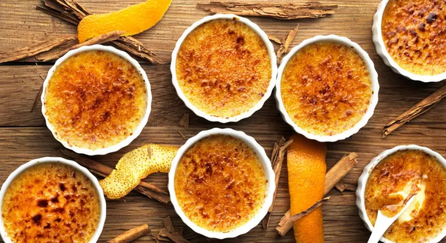 Flera krämiga crema catalana i vita skålar, toppade med karamelliserat socker. Apelsinskal och kanelstänger ger en härlig doft.