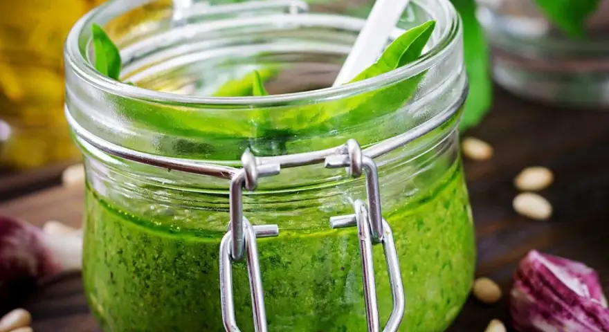 Un pot de pesto ail des ours, vert vif, avec une cuillère blanche. Des pignons de pin et de l'ail parsèment la table en bois.