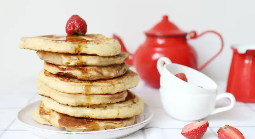 Pile de pancake sans gluten, nappés de sirop et garnis d'une fraise. Une théière rouge et des tasses complètent la scène.