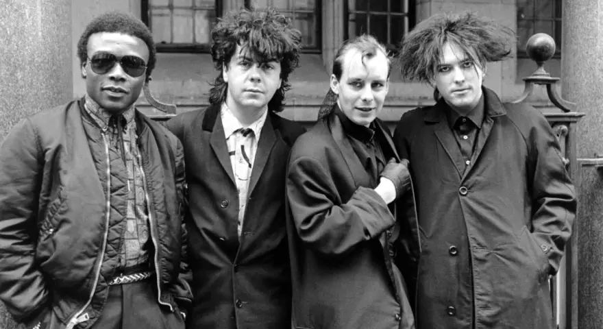 Vier Männer in dunkler Kleidung, Post-Punk-Ästhetik, stehen vor einem Gebäude.