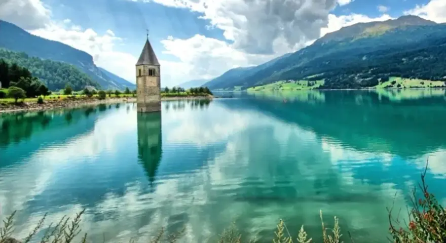Il lago del campanile, con la sua torre sommersa, riflette montagne verdi e un cielo nuvoloso.