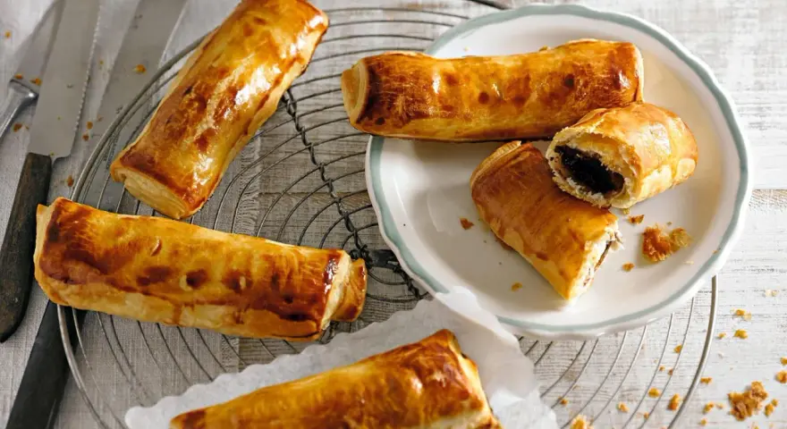 Goldbraune, knusprige Pain au Chocolat auf einem Gitter, einige auf einem Teller, einer aufgeschnitten.