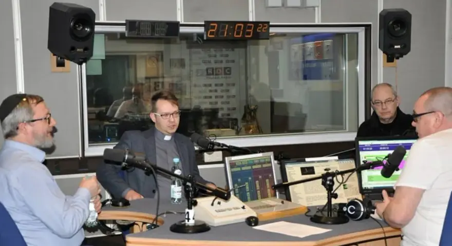 W studiu radiowym czterech mężczyzn, w tym ksiądz i osoba w jarmułce, rozmawia przy mikrofonach. W tle widać zegar i logo RDC.