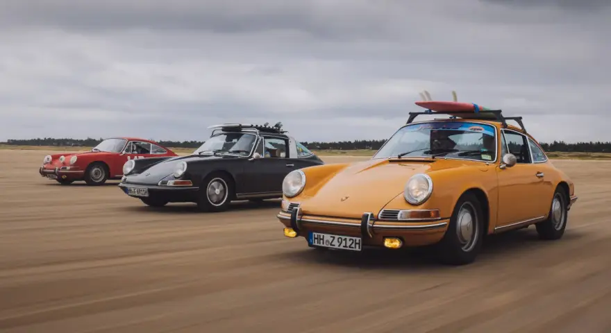 Trzy klasyczne Porsche jadące po plaży. Zdjecie w ruchu, z deskami surfingowymi na dachu jednego z nich.