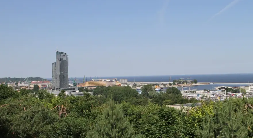 Widok na port w Gdyni z dominującym wieżowcem. W oddali morze, a na pierwszym planie zieleń drzew. Kamienna Góra w Gdyni oferuje takie panoramy.