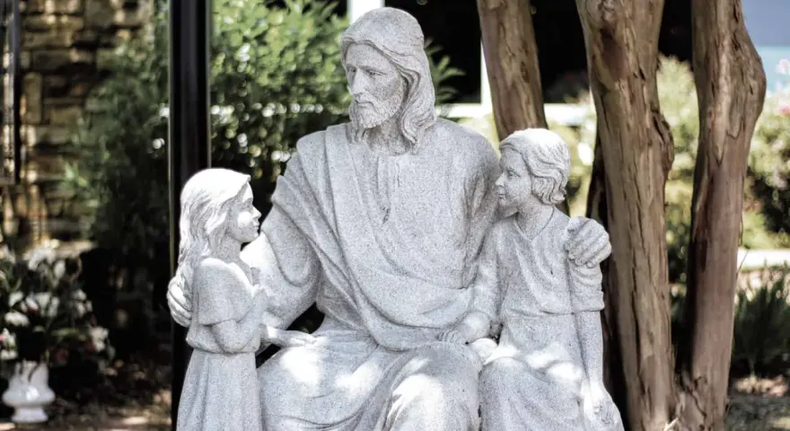 Steinfigur von Jesus, der zwei Kinder umarmt. Er wirkt wie ein liebevoller Gottvater, der seine Schützlinge beschützt.