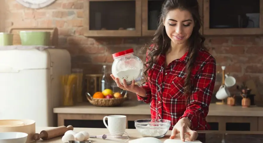 Junge Frau liest Rezept und hält Mehl bereit. Sie überlegt, ob sie Wasser statt Milch beim Backen verwenden soll.