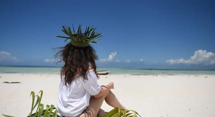 Kobieta w koronie z liści siedzi na plaży, marząc o tym, jak dolecieć na Zanzibar. Błękitne niebo i turkusowa woda zapraszają do podróży.