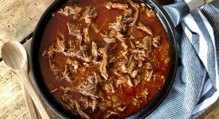 En gjutjärnspanna fylld med saftig, långkokt pulled beef i en mustig sås. Två träskedar ligger bredvid, redo att servera.