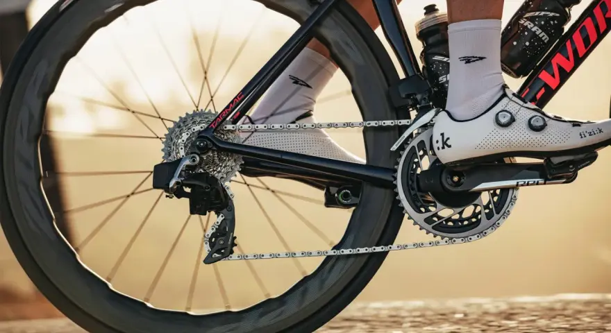 Close-up of a cyclist's shoe and drivetrain, showing the chain, gears, and crankset. The focus is on the power transfer, hinting at the cycling watts being generated.