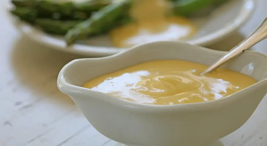 Krämig hollandaisesås i ett serveringskärl, perfekt för ditt hollandaisesås recept. Sparris syns i bakgrunden.