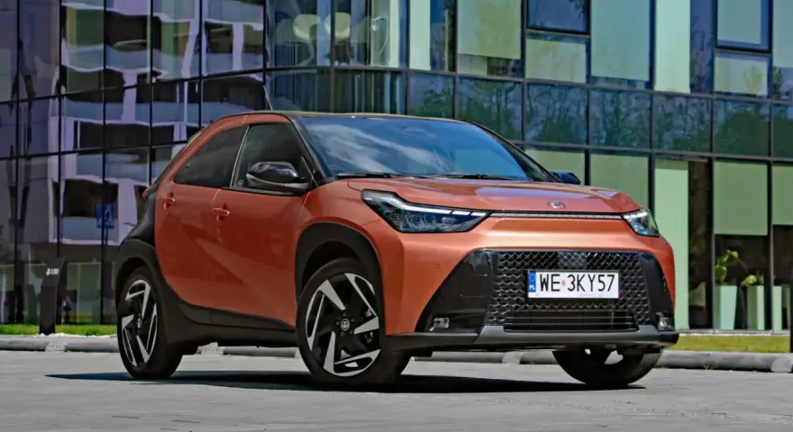 Pomarańczowy Toyota Aygo X Hybrid z czarnymi detalami stoi przed nowoczesnym budynkiem.