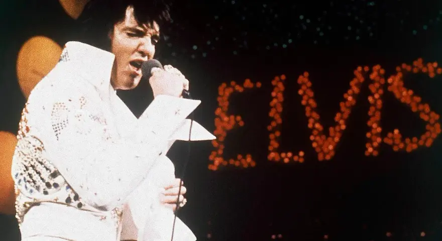 Elvis Presley singt in seinem berühmten weißen Anzug in Las Vegas, während sein Name in roten Lichtern erstrahlt.
