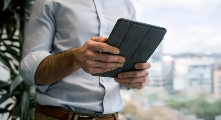 Mężczyzna w jasnej koszuli i brązowych spodniach trzyma tablet w szarej okładce. e cmr.