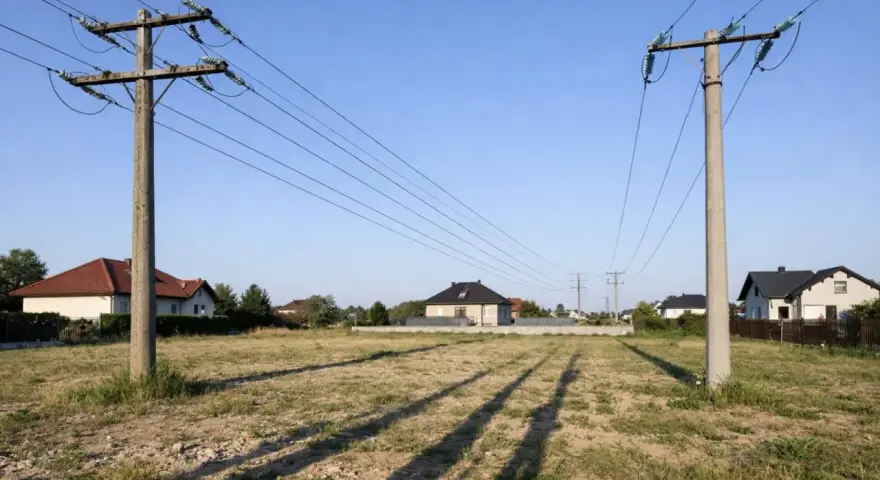 Linia wysokiego napięcia nad działką z rozpoczętą budową domu. Słupy energetyczne i przewody przecinają krajobraz z kilkoma domami w tle.