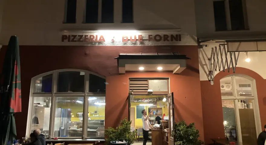Abendstimmung vor der Pizzeria "Due Forni". Ein Mann steht im Eingangsbereich, während Gäste an Tischen im Freien sitzen.