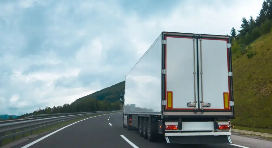 Ciężarówka na autostradzie, z tyłu widoczne białe drzwi naczepy. W tle góry i chmury. Tablice wyróżniające przepisy są częścią oznakowania pojazdu.