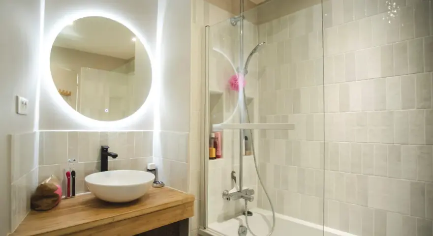Une **salle de bain blanche** moderne avec un lavabo rond sur un meuble en bois, un miroir lumineux et une douche avec carrelage clair.