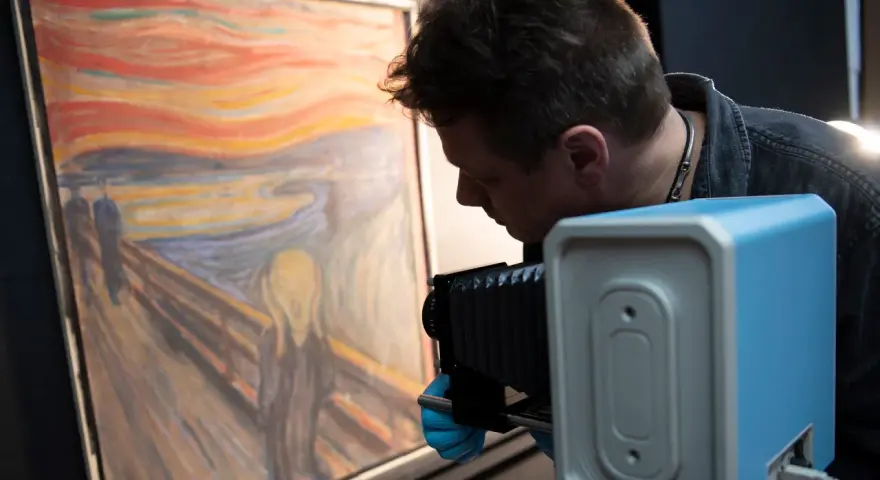 En man med blå handskar fotograferar "Skriet". Vem har målat skriet? Edvard Munch.