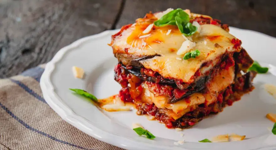 Una fetta di parmigiana di melanzane al forno, stratificata con melanzane, sugo e formaggio filante, guarnita con basilico fresco.