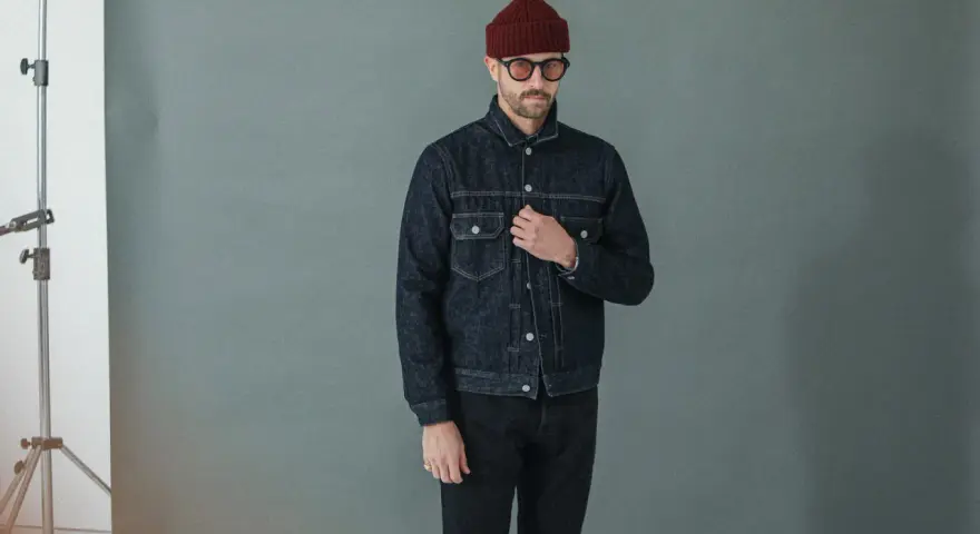 Homme portant une veste en jean sombre et un pantalon noir, bonnet bordeaux et lunettes rondes.