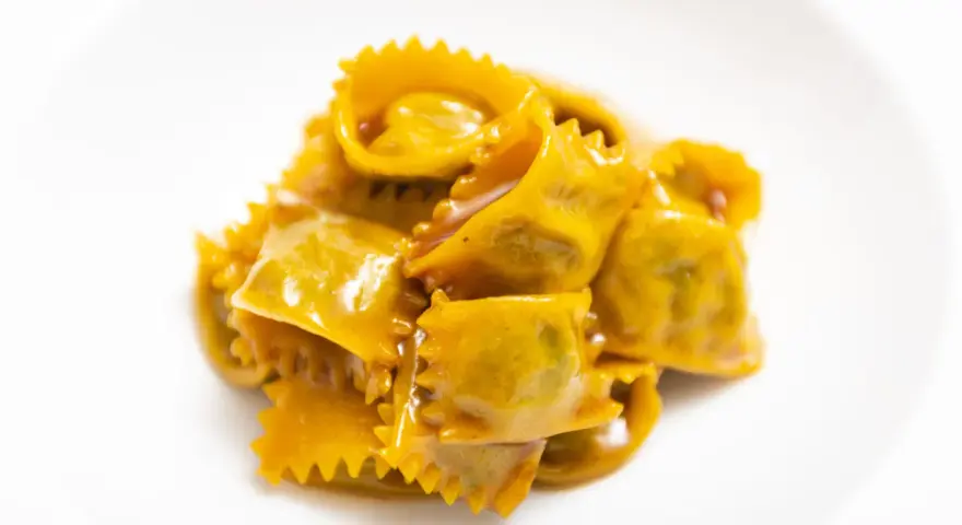 Un mucchietto di ravioli del plin, dorati e conditi con un sugo cremoso, su un piatto bianco.