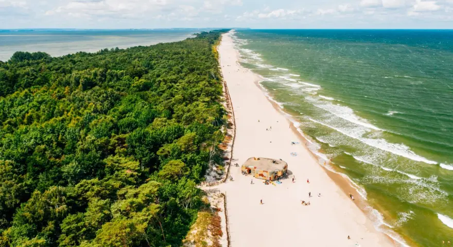 Majówka nad morzem: zielony las, piaszczysta plaża z kilkoma osobami i bunkrem w kamuflażu, szmaragdowe fale Bałtyku.