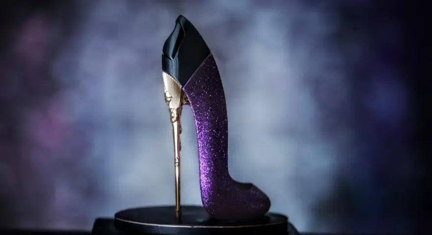 Elegancki perfumy bucik w kolorze fioletowym, ozdobiony złotymi detalami i brokatem, stoi na czarnym postumencie.