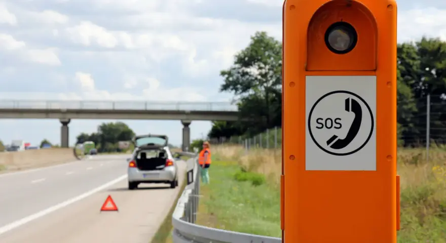 Ihr Fahrzeug ist auf der Autobahn liegen geblieben. Eine Frau in Warnweste steht neben dem offenen Kofferraum, ein Warndreieck liegt auf der Fahrbahn. Eine Notrufsäule steht im Vordergrund.